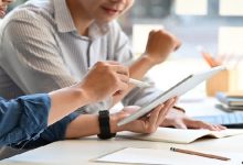 Cropped image of smart men working as project manager discussing/talking about their project by using a computer tablet while sitting together at the long white desk over glass wall as background.